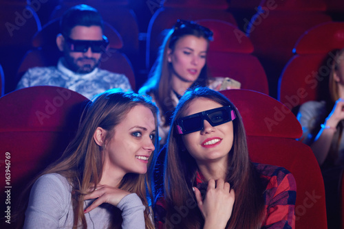 Attractive young women watching a movie together. Female friends chatting while enjoying a film premiere at the movie theatre entertaining technology 3D viewer spectators audience activity friendship.