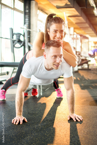 Young sporty couple exercising together in gym. Man doing push ups while girl lying on him.