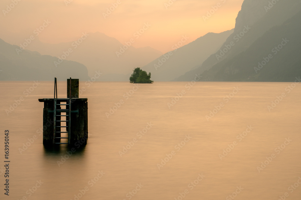 hazy evening with silhouette of mountains in the background at a lake in Switzerland with a diving platform and an island in the center hazy evening with silhouette of mountains in the background at a lake in Switzerland with a diving platform and an island in the center
