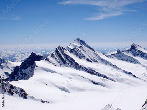 two mountain climbers on a narrow and exposed snowy summit ridge with a spectacular view of the Swiss Alps