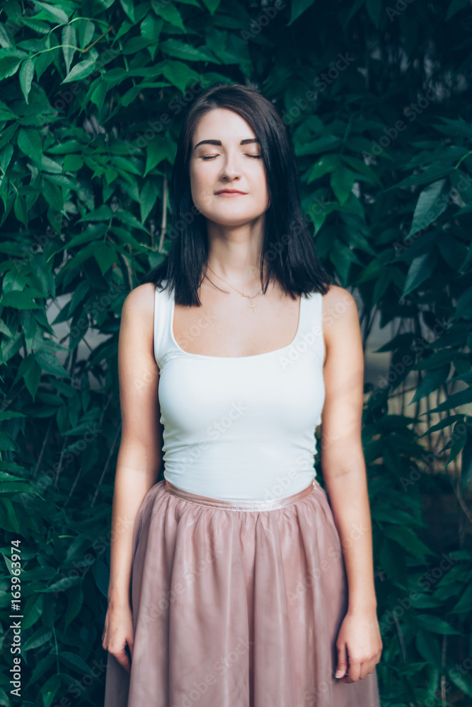 Portrait of a young, beautiful brunette who stands near a green tree foliage with closed eyes Portrait of a young, beautiful brunette who stands near a green tree foliage with closed eyes