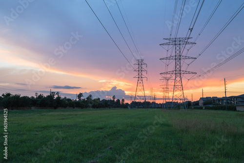 Group silhouette transmission towers (steel lattice/power tower, electricity pylon) next to apartment complex at sunset in US. Texture high voltage pillar, overhead power line, industrial background.