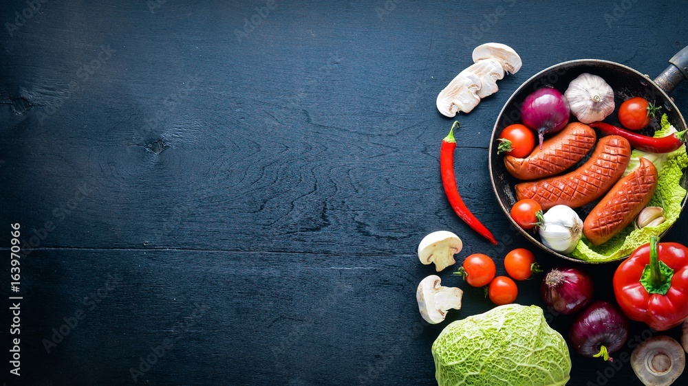 Grilled sausages in a pan with fresh vegetables. Wooden on the black surface. Top view.