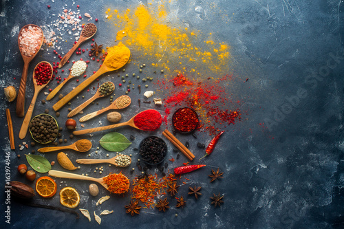 Various indian spices in wooden and silver spoons and metal bowls, seeds, herbs and nuts on dark stone table. Colorful spices, top view,  space for text. Organic food, healthy lifestyle.