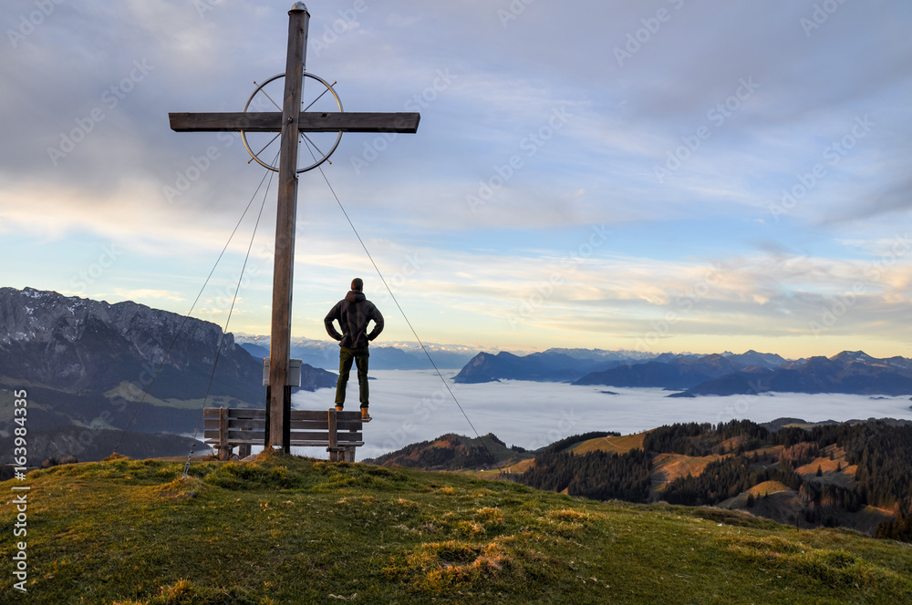 Obraz premium Lonely hiker standing at a summit cross at Wandberg mountain in Chiemgau Alps mountain range in Austria shortly after sunrise. Tranquil scene with fog in the lowland. The cross is a christian symbol.