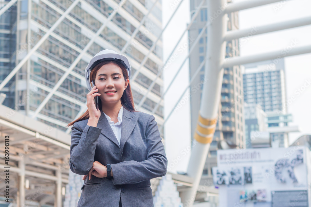 Portrait of beautiful young engineer woman wear a white safety helmet. She are smile and talking on smartphone with commitment to success with building background