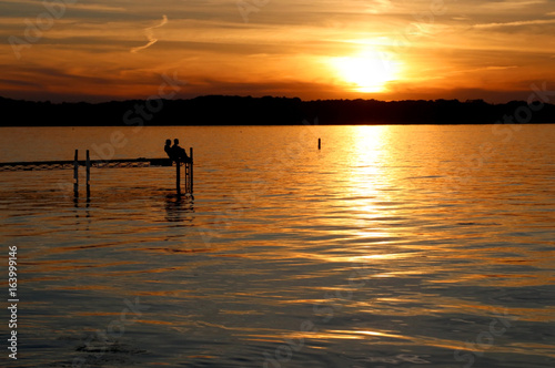 Summer sunset over the lake. Landscape with golden sunset and silhouettes of people enjoying the beautiful evening on a lake Mendota pier in the city of Madison, Wisconsin, USA. © Maryna