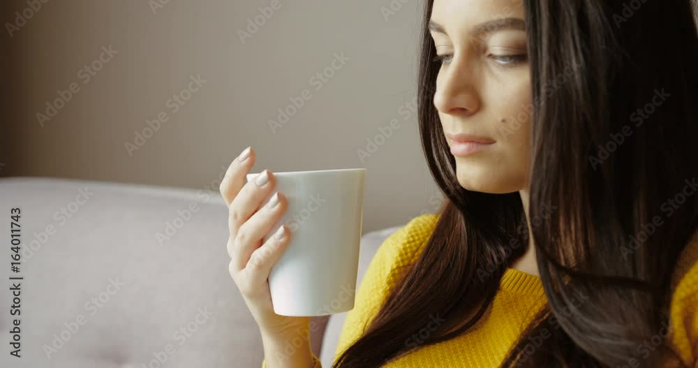 Attractive young woman sitting on the couch, drinking coffee, tea and using smart phone. Chatting, surfing on the internet. Close up. Slow motion Attractive young woman sitting on the couch, drinking coffee, tea and using smart phone. Chatting, surfing on the internet. Close up. Slow motion