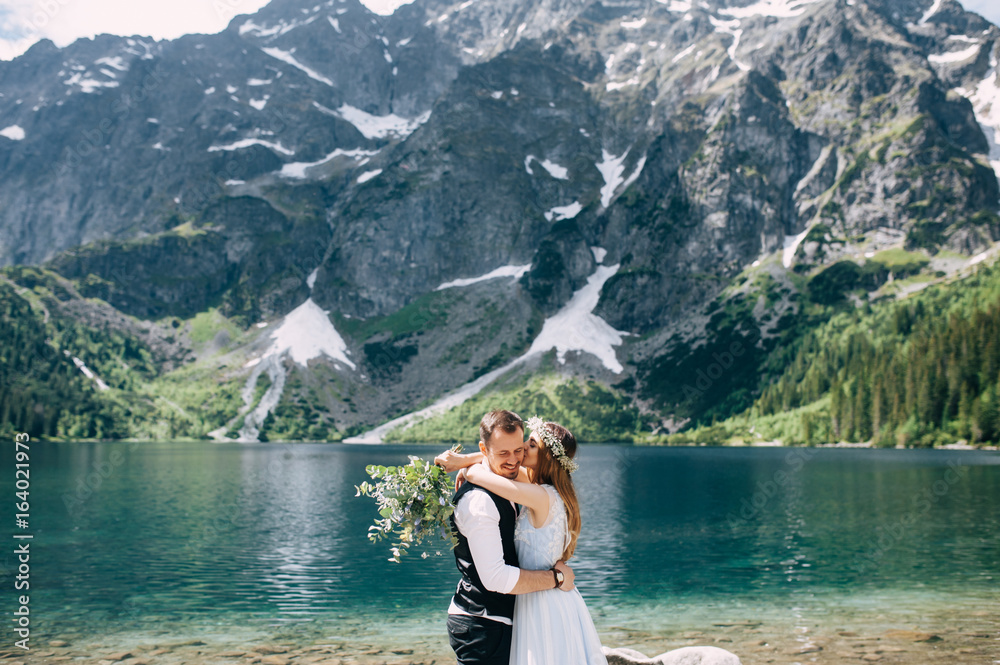 bride with the beautiful blue dress and groom hugging with views of the beautiful green mountains and lake with blue water