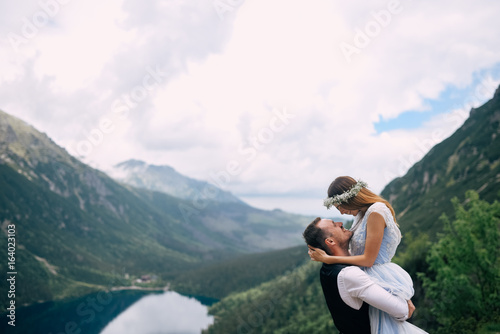 bride with the beautiful blue dress and groom hugging with views of the beautiful green mountains and lake with blue water