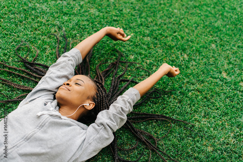 Принти на полотні Delighted afro-american teenager is enjoying music while laying on the grass and raising hands