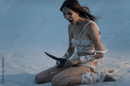 Quadro su tela Girl in image of Egyptian mummy sits on sand with  metal chain and knife in her hands and screams in fury