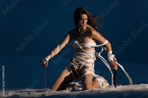 Quadro em tela Girl in image of Egyptian mummy kneels on sand with metal chain and knife in her hands