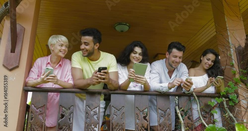 Young Group Of Friends Using Modern Gadgets Messaging Stnd On Hotel Balcony, Mix Race People Chatting Online Slow Motion 60 photo