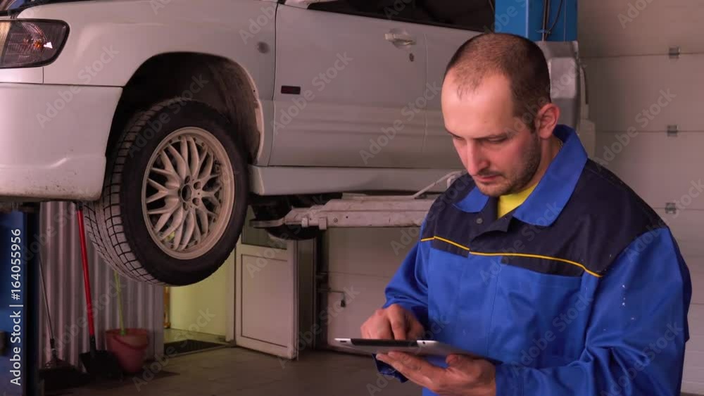 Car mechanic examines the results of electronic car diagnostics with tablet in front of the car on lift.