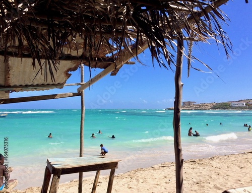 Sunny summer afternoon at beautiful Hellshire (Healthshire) Beach Portmore, Jamaica (St. Catherine Parish) near Kingston: turquoise ocean, white sand beach & blue sky, beach hut, Jamaicans swimming