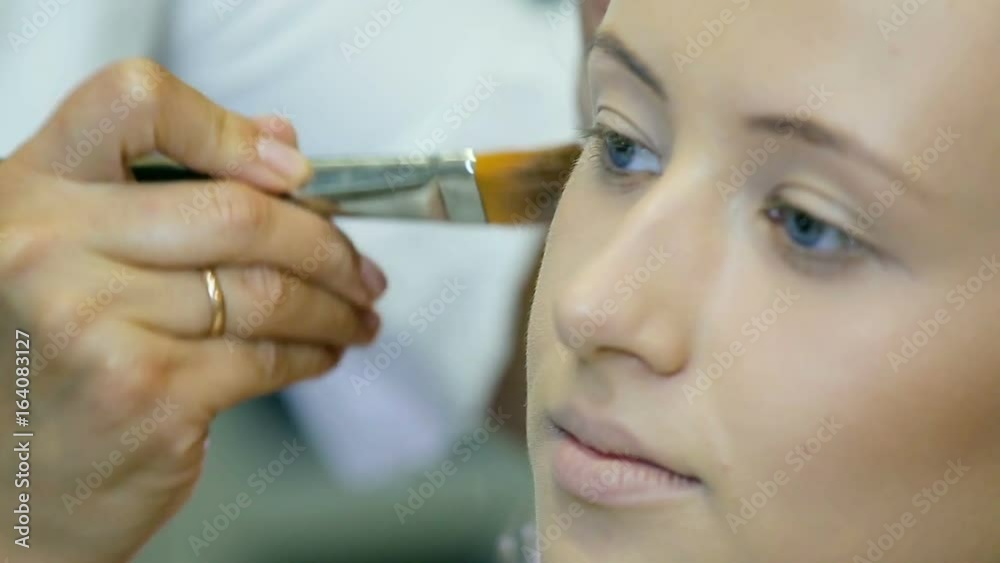 Professional make-up artist applying tonal cream to blond long curl hair and blue eyes model skin. Shaping face. Professional make-up artist applying tonal cream to blond long curl hair and blue eyes model skin. Shaping face.