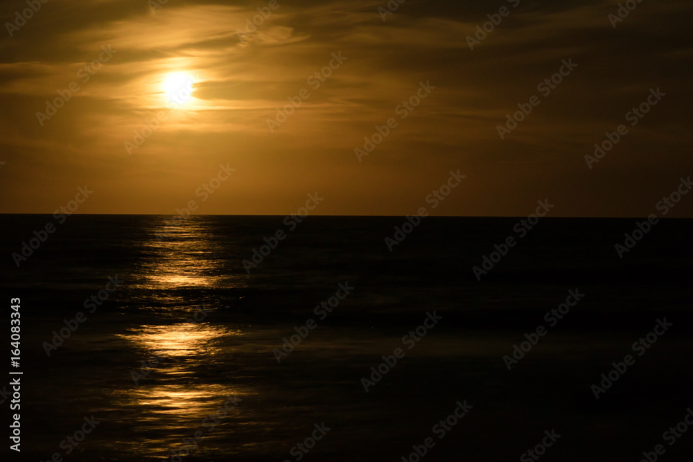 full moon rises over the ocean with a soft orange reflection on the water and the incoming waves