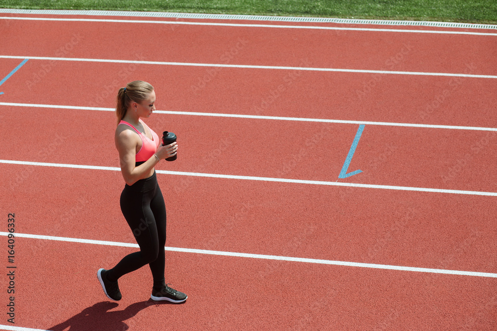 Young sporty woman athlete in sportswear drinking water or protein cocktail from shake on stadium track, workout concept