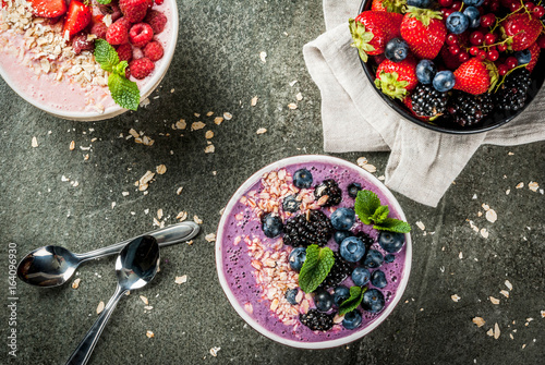 Ideas for healthy summer breakfast. Smoothies in bowls wth red (strawberry raspberry currant), blue (blueberry blackberries) berry With oatmeal, fresh berries. Dark stone table. Top view copy space