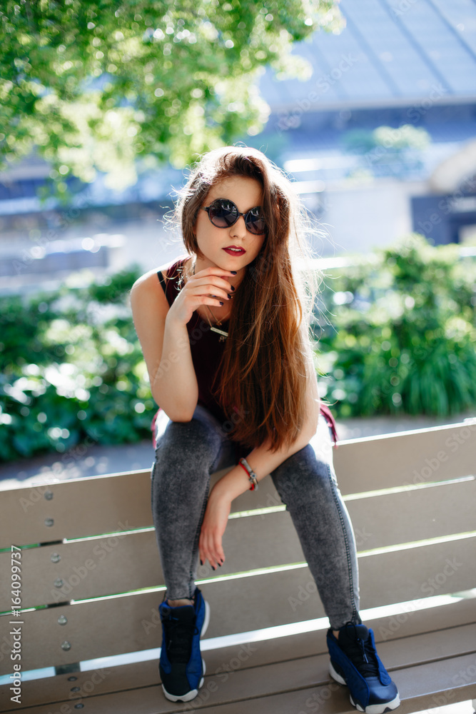 Portrait of beautiful sexy smiling white Caucasian girl woman with long blond red hair, wearing jeans and t-shirt, sitting on bench in park outside with sunglasses. Hot summer day, lifestyle concept.