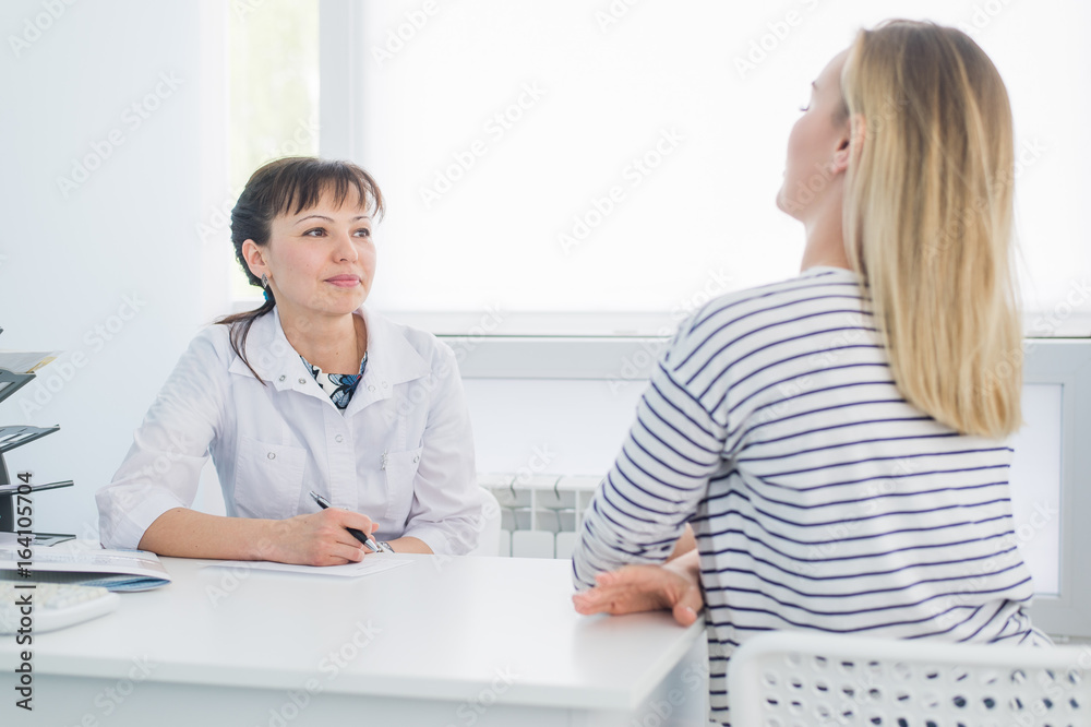 Smiling patient receiving a medical consultation and looking at camera, the female doctor is sitting at desk on the background Smiling patient receiving a medical consultation and looking at camera, the female doctor is sitting at desk on the background