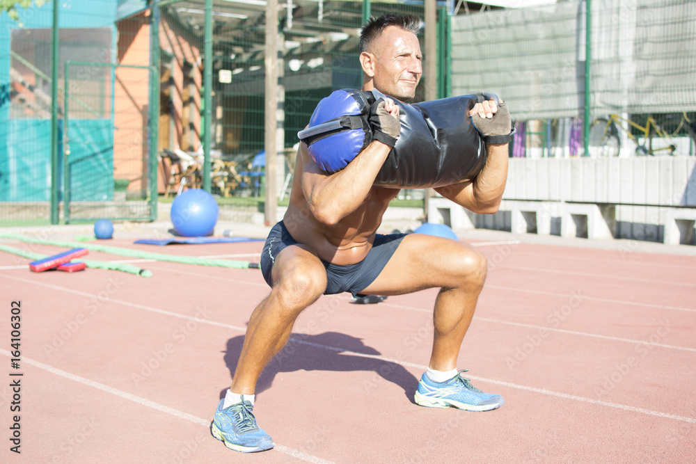 Handsome middle aged man working out on a running track. Healthy adult man doing squats with a heavy bag on the track. Tanned skin and shirtless middle-aged man.
