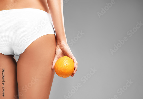 Young woman holding orange on grey background. Cellulite problem concept