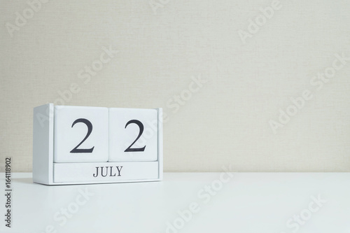 Closeup white wooden calendar with black 22 july word on blurred white wood desk and cream color wallpaper in room textured background with copy space , selective focus at the calendar