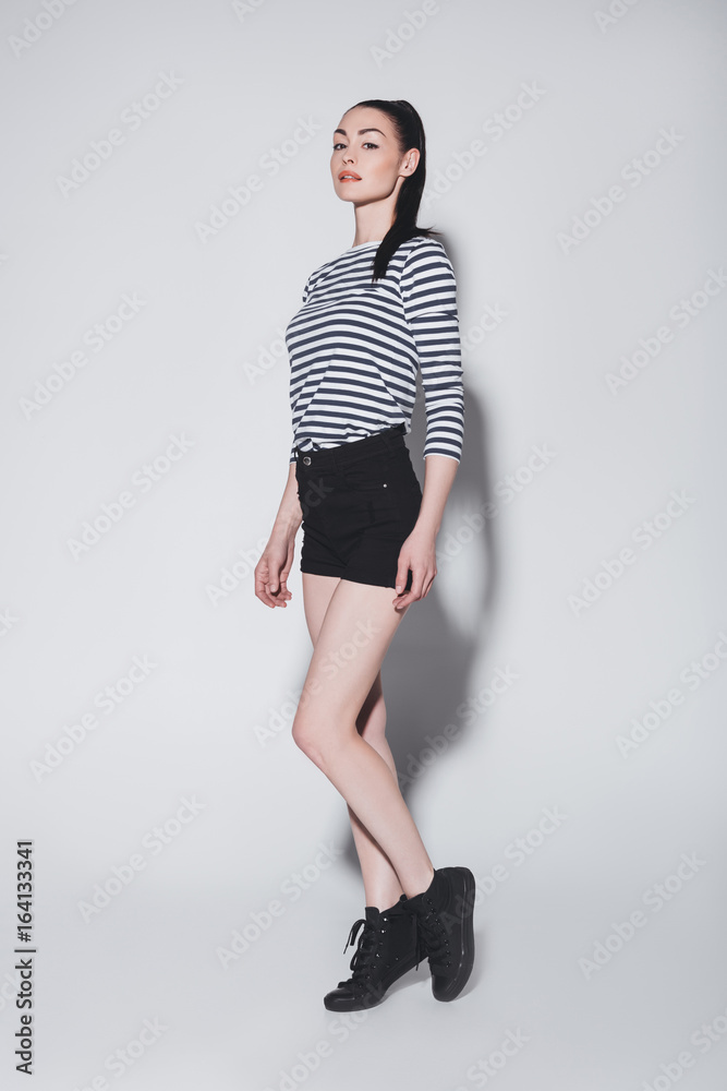 Beautiful stylish young woman in shorts posing and looking at camera on grey