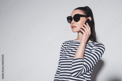 Gorgeous stylish young woman in sunglasses talking on smartphone isolated on grey