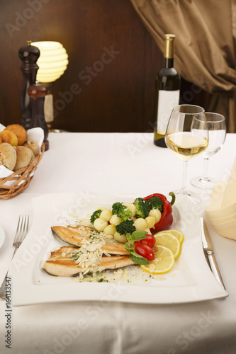 Hake fillet with grilled vegetables arranged on a plate, Wine bottle and wineglass in background, Traditional dish in elegant setting, Selective focus with soft light