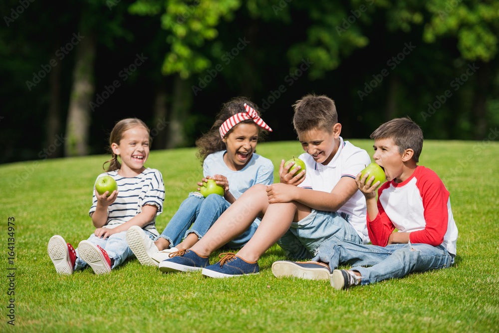Happy multiethnic children eating green apples while sitting together on green grass Happy multiethnic children eating green apples while sitting together on green grass