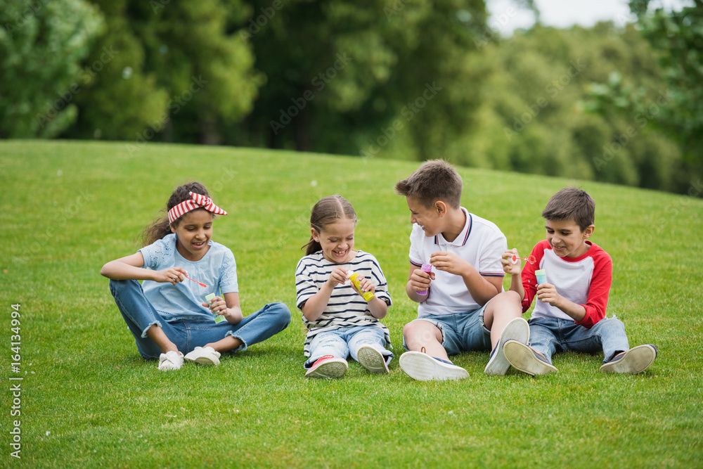 Fototapeta premium Happy multiethnic kids playing with soap bubbles while sitting on green meadow in park