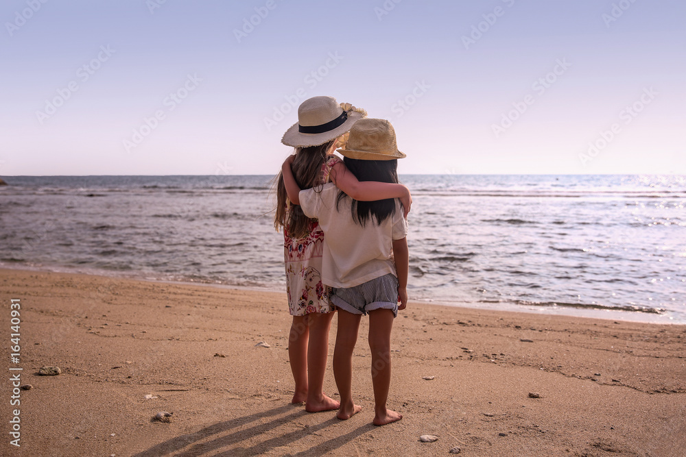 Two child girls stand together on the beach and looking at sea. Sunny day. Back view. Holiday concept
