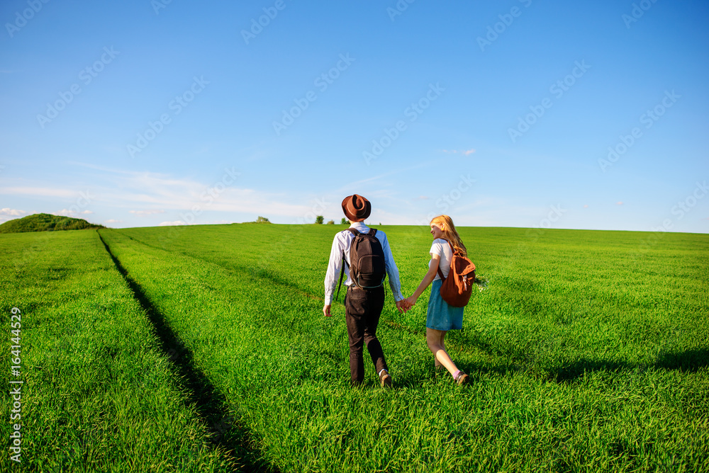 With a backpack, a man in a hat and a woman with long hair go along the path. A couple walks along the meadow With a backpack, a man in a hat and a woman with long hair go along the path. A couple walks along the meadow