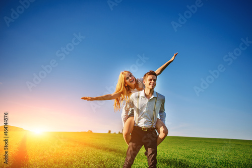 Smiling Man is holding on his back happy woman, who pulls out her arms and simulates a flight against the background of the blue sky and the green field