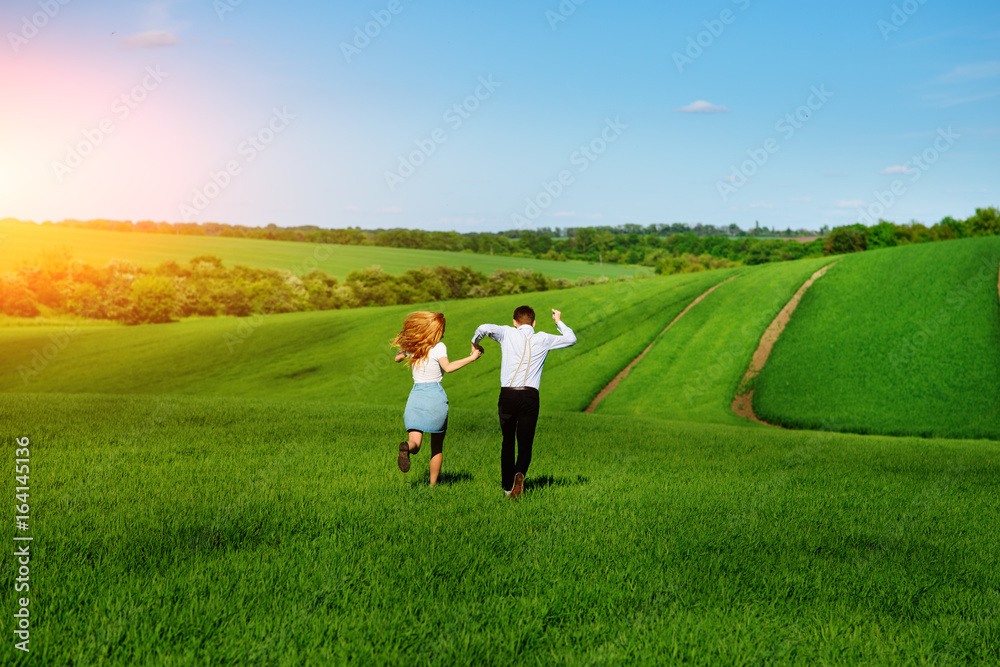 Young happy lovers running on meadow with green grass and blue sky, From the back Young happy lovers running on meadow with green grass and blue sky, From the back