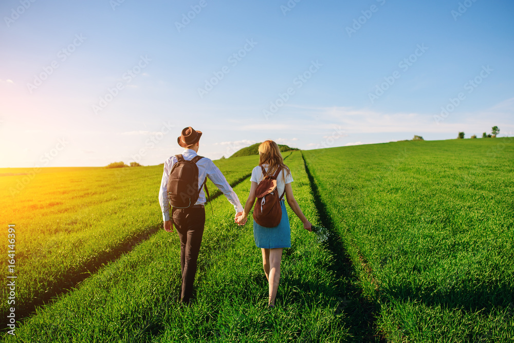 With a backpack, a man in a hat and a woman with long hair go along the path. A couple walks along the meadow With a backpack, a man in a hat and a woman with long hair go along the path. A couple walks along the meadow