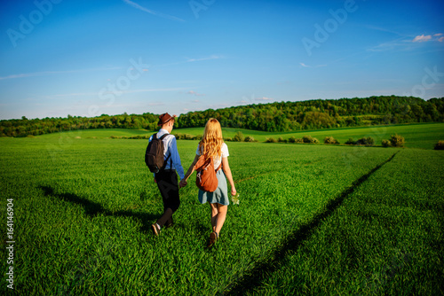 With a backpack, a man in a hat and a woman with long hair go along the path. A couple walks along the meadow