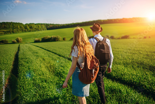 With a backpack, a man in a hat and a woman with long hair go along the path. A couple walks along the meadow