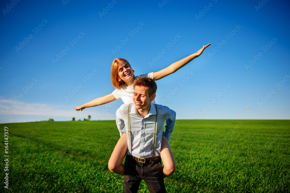 Smiling Man is holding on his back happy woman, who pulls out her arms and simulates a flight against the background of the blue sky and the green field Smiling Man is holding on his back happy woman, who pulls out her arms and simulates a flight against the background of the blue sky and the green field