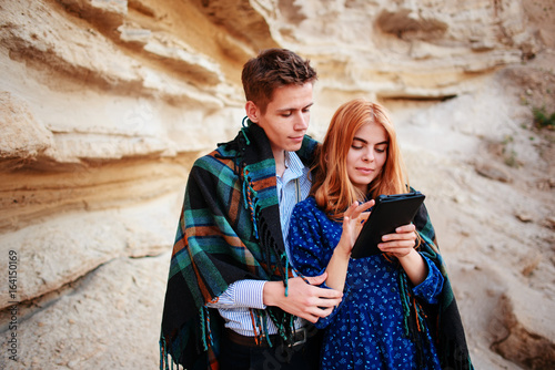 Beautiful woman and handsome man wrapped in a blanket. They are smiling and looking at the screen of a tablet on the background of a sand quarry © cezarksv