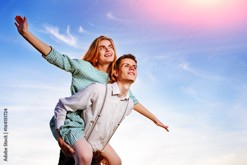 Young guy piggybacking cheerful girlfriend like airplane on the background of sky Young guy piggybacking cheerful girlfriend like airplane on the background of sky