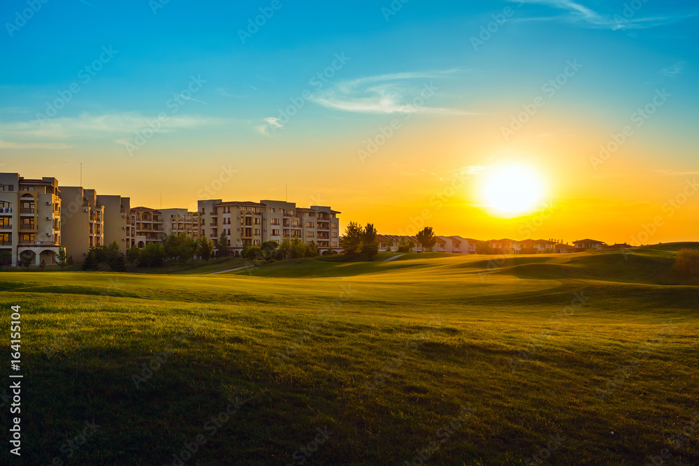 Fototapeta premium Green field for golf near apartment buildings, in dusk at sunset, tranquil place for rest