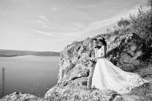 Fantastic wedding couple standing on the edge of rocky precipice with a perfect view of lake on the background. Black and white photo.