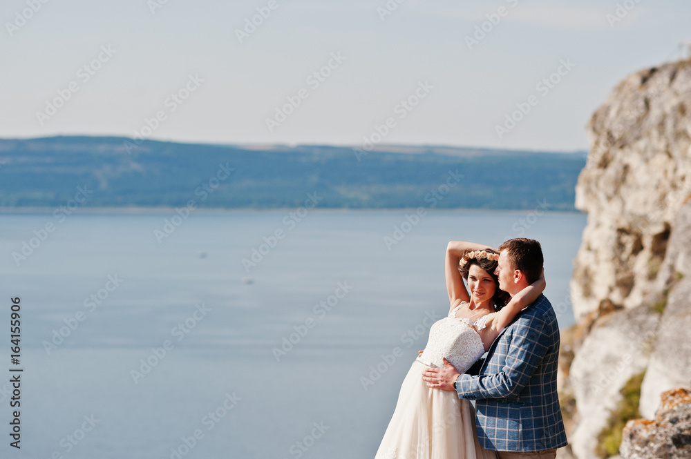 Fantastic wedding couple standing on the edge of rocky precipice with a perfect view of lake on the background.