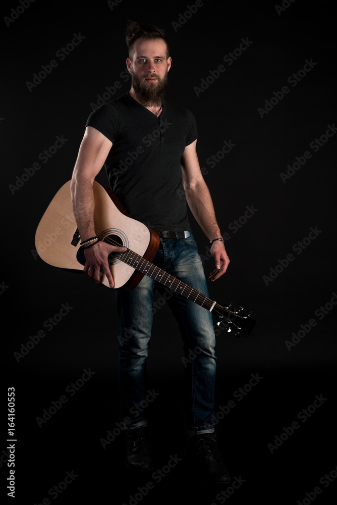 Fototapeta premium A charismatic and stylish man with a beard stands full-length with an acoustic guitar on a black isolated background. Vertical frame