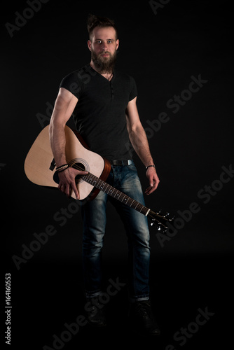 A charismatic and stylish man with a beard stands full-length with an acoustic guitar on a black isolated background. Vertical frame