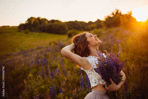 Golden rays of evening sun illuminate gorgeous lady with bouquet on the field